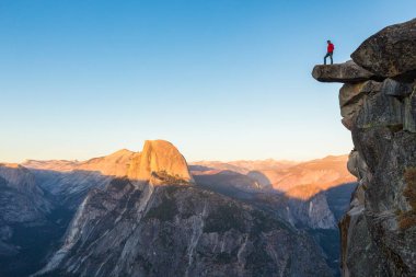 A fearless hiker is standing on an overhanging rock enjoying the view towards famous Half Dome at Glacier Point overlook in beautiful evening twilight, Yosemite National Park, California, USA clipart