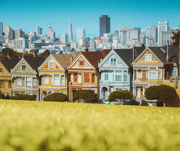 Classic postcard view of famous Painted Ladies, a row of colorful Victorian houses located at Alamo Square, with the skyline of San Francisco in the background