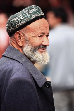 Hotan, Çin-3 Ekim 2017: Uygur Türklerinin sakallı yaşlı adamı, şehir pazarında etnik kimliğinin bir göstergesi olarak yeşil-beyaz doppa-kafatası kapağı takıyor. Xinjiang Bölgesi.