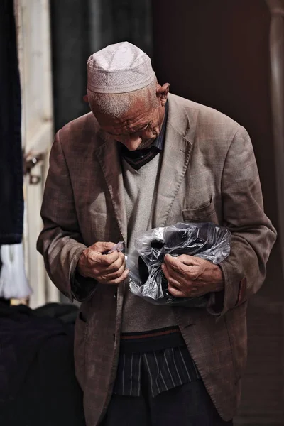 中国和田 2017年10月3日 维吾尔族的老人头戴白色圆帽 头戴一顶白色圆帽 小心翼翼地在一家塑料酒吧里购买他在城市集市上买的东西 新疆地区 — 图库照片