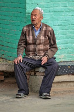 Hotan, Xinjiang, Çin-3 Ekim 2017: Kareli kahverengi hırka ve beyaz doppa şapkalı Uygur halkının yaşlı adamı, pazar günü pazarın arka tarafındaki yeşil duvara bağlı bir bankta oturuyor.
