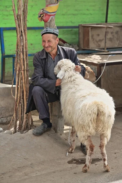 Hotan, China-October 4, 2017: Uyghurs are a Turkic people living mainly in the Xinjiang Uyghur Autonomous region. Smiling livestock handler sitting at the Livestock Market with his fat-tailed sheep.