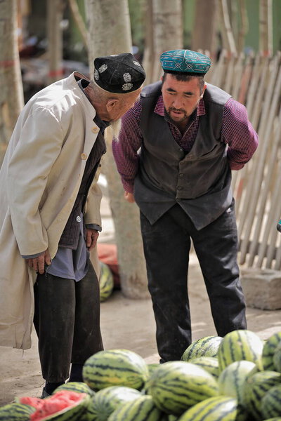 Hotan, China-October 4, 2017: Uyghurs are a Turkic people living mainly in the Xinjiang Uyghur Autonomous region. Livestock Market scene: attentive watermelon seller and white overcoat-dresssed buyer.