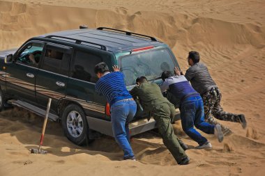 Keriya County, China-October 5, 2017: Car-pushing drivers unstuck a green off-road car for touristic service stuck in sand while driving through the treacherous loose dunes of the Taklamakan desert. clipart