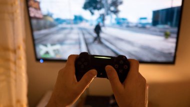 Video oyunu oynayan adam. Eller konsol kontrolörünü tutuyor. Televizyonda futbol ya da futbol maçı. Geniş ekran televizyon klozetin üzerinde duruyor