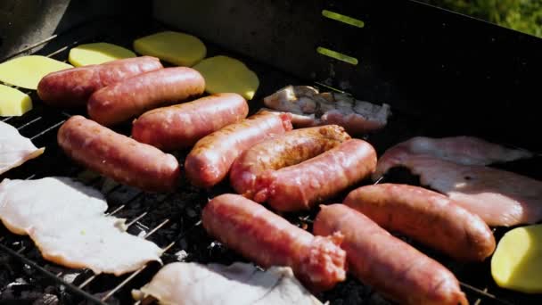 바베큐에서 손잡이로 맛있는 감자와 쥬스 소시지를 굽는 거. 검은 바베큐그릴 위에서 딱딱 한 감자와 소시지를 손으로 금속 통의 도움을 받아 굽는다 — 비디오