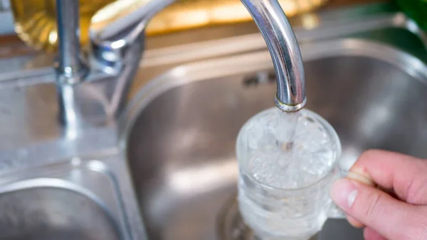 Male hand with a glass under the tap. Glass in male hand is filled with clean transparent water under the tap with metal sink on the background.