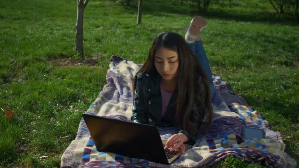 Стильная девушка лежит на лужайке и печатает на ноутбуке в парке — стоковое видео