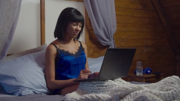 オンラインビデオ通話のためのラップトップを使用してベッドの上でリラックス青いパジャマの正のエレガントな若いアフリカ系アメリカ人女性 豪華な多民族の女性でベッドでビデオチャットのためのコンピュータウェブカメラを使用して夜 — ストック動画