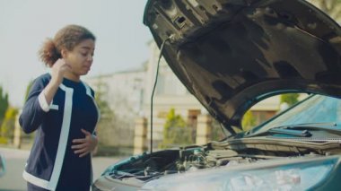 Kafası karışmış bir kadın sokakta bozuk bir araba motoruna bakıyor.