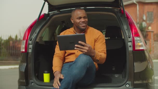 Homem relaxado usando tablet pc enquanto sentado no porta-malas do carro — Vídeo de Stock