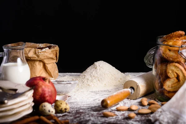 Vista de cerca de deliciosas galletas caseras en botella de vidrio e ingredientes para hacer masa en la mesa sobre negro - foto de stock