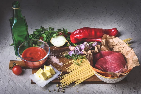 Vista de perto de carne crua, espaguete não cozido, pimenta, cebola, manteiga, molho de tomate e garrafa de óleo na tábua de corte — Fotografia de Stock