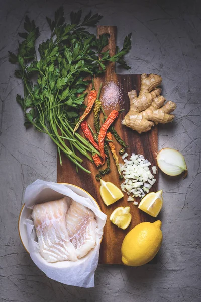 Top view of raw fish in bowl, parsley, peppers, salt, onion, lemon and ginger on wooden cutting board — Stock Photo