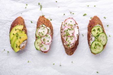 Healthy sandwiches with grain bread, ricotta, avocado, cucumber, radish and mustard micro greens. On white paper, top view. Healthy snack