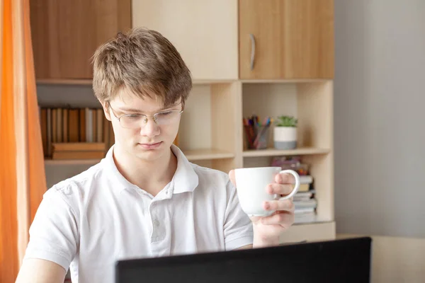 Student in glasses learning at home using laptop. Young man holding mug. Home school, online education, home education - Image