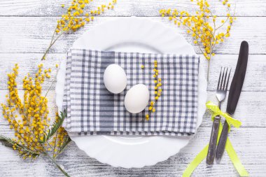 Paskalya sofrası. Beyaz yumurta, keten peçete, beyaz tabak, mimoza çiçekleri ve ahşap bir masada çatal bıçak takımı. Üst görünüm
