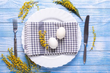Paskalya sofrası. Beyaz yumurta, tabakta peçete, mimoza çiçekleri, çatal, mavi ahşap bir masada bıçak. Yukarıdan görüntüle