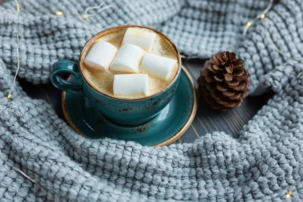 Cup of coffee, marshmallow, warm knitted sweater on wooden background. Warm lights. Cozy winter morning. Lifestyle concept. Selective focus.
