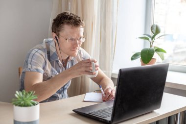 Kulaklıklı programcı evden çalışıyor, elinde kupa var. Gözlüklü bir adam monitöre bakıyor. Uzak çalışma konsepti - Resim