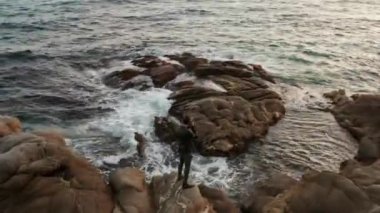 Siyah elbiseli bir adam bir kayanın üzerinde duruyor ve bir kamerayla denizin fotoğraflarını çekiyor. İHA kullanarak yukarıdan ateş etmek