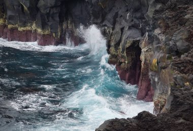 Büyük güçlü mavi okyanus dalgaları siyah kaya kıyısına vuruyor. Yüksek su damlası ve beyaz deniz köpüğü