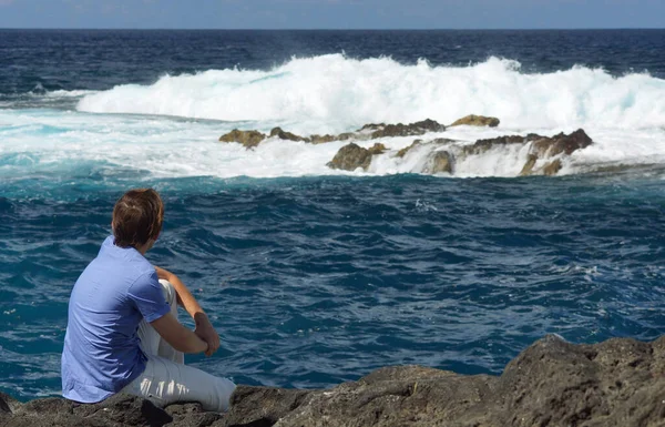 Young man sitting on rock shore and looking at ocean. View from behind. Blue ocean with high waves, water splash — Stock Photo, Image