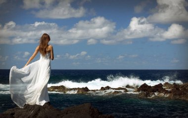 Açık beyaz gelinlikli genç gelin deniz taşları kıyısında duruyor ve okyanusa bakıyor. Arka plan