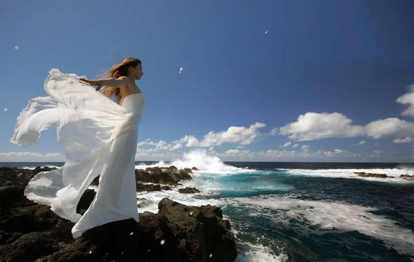 Young bride with white wings of wedding dress on rock sea shore. Side view. Ocean waves, splash of water