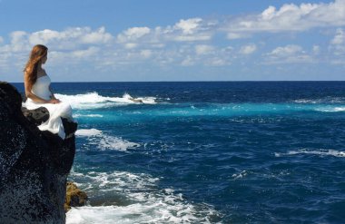 Gelinlikli gelin okyanus kıyısında oturmuş okyanusa bakıyor. Okyanusta düğün töreni