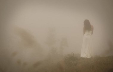 Woman in long white dress in foggy misty landscape. Blur discernible outline of slim figure. View from behind. Mistery