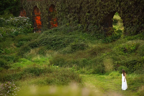 Young bride in white dress standing in field near grassy brickwall. Small woman figure in green fairy tale landscape — 스톡 사진