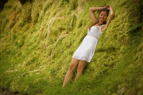 Beutiful young girl leaning against green moss wall near road. Expression of relaxation and happiness on face — 스톡 사진