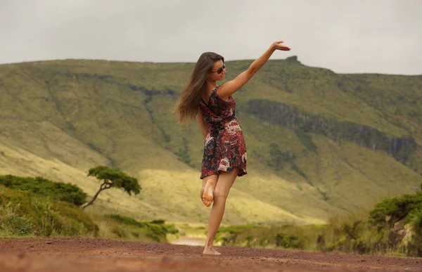 Young woman in dress turning head backwards with raised hand on country road. View from behind. Good bye hand in the air