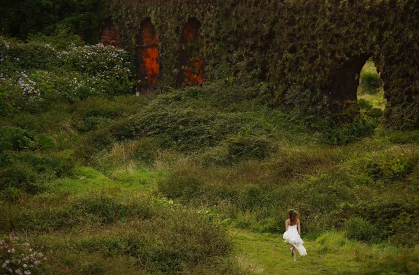 Young bride in white dress running barefoot in field between bushes. Small figure in green fairy tale landscape