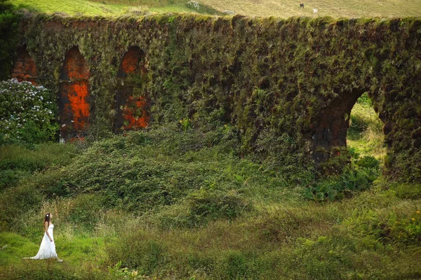 Bride in white dress standing near grassy brickwall in green field. Small woman figure in green fairy tale landscape