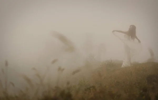 Woman in long white dress in foggy misty landscape. Blur discernible outline of slim figure. View from side. Mistery