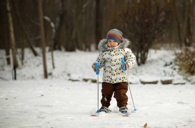 Küçük kayakçı parlak giysiler içinde elinde sopalarla küçük bir kayağın üzerinde duruyor. Karlı kış parkının arka planı