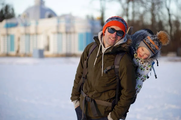 Hombre sonriente retrato con niño pequeño en mochila portabebés. Sonriente familia feliz. Fondo del parque de invierno nevado — Foto de Stock