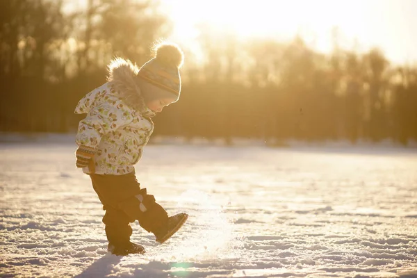 Маленький мальчик в яркой одежде, шляпе с помпоном и варежками, стоящий на солнце и играющий со снегом в солнечном снежном парке — стоковое фото