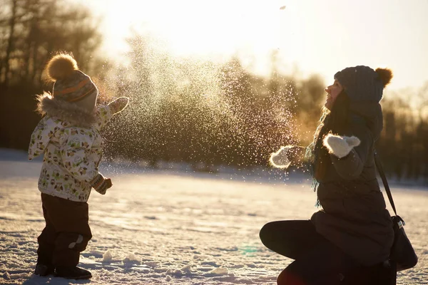 Mujer joven con hijo pequeño vomitando nieve en el fondo del soleado parque de invierno, foto contra el sol. Familia feliz — Foto de Stock