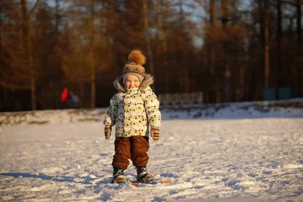 Pequeno esquiador menino sorridente em roupas brilhantes e chapéu com pompom de pé no esqui no fundo do ensolarado parque de inverno nevado — Fotografia de Stock