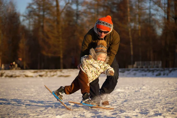 Молодой человек учит своего маленького ребенка кататься на лыжах. Улыбающийся мальчик падает. Солнечное фото на фоне зимнего снежного парка — стоковое фото