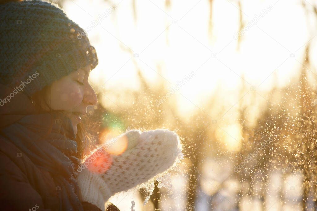 Side portrait of young woman in woolen hat and white woolen mittens throwing up snow at sunset time, closeup