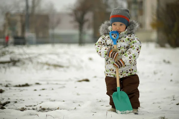 Портрет маленького мальчика с вдумчивым взглядом с большой лопатой в руке, стоящей в снегу. Фон снежного зимнего парка — стоковое фото