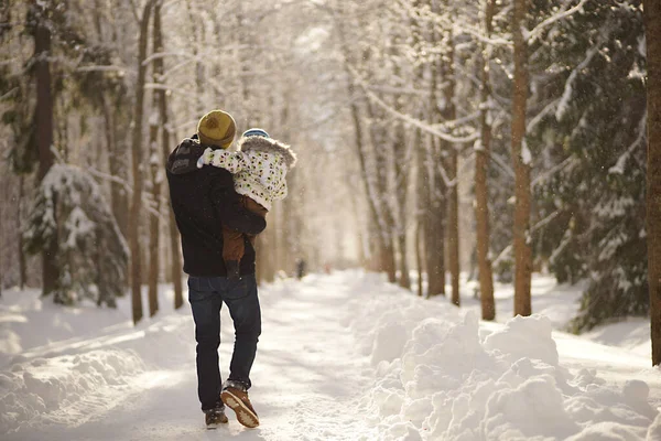 손에 아이를 안고 햇빛 이 잘 드는 겨울 공원, 눈덮인 나무와 눈 언덕을 걷고 있는 젊은이. 뒤에서 본 모습 — 스톡 사진