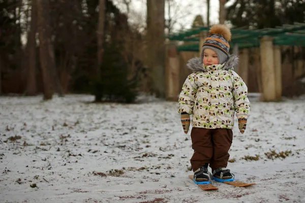 Little boy skier in bright clothes standing on small ski on snow. Background of snowy winter park — Stock Photo, Image