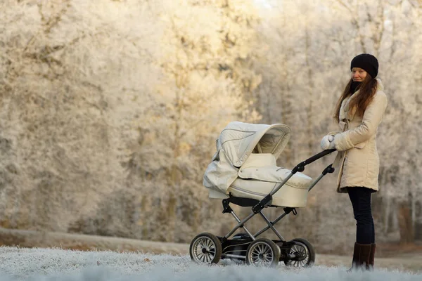 Молодая женщина в белом халате стоит с белой детской коляской в солнечном зимнем парке. Уход за детьми и любовь — стоковое фото