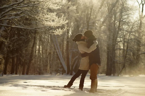 Pareja joven abrazándose y besándose en el soleado bosque nevado, fondo de árboles helados, amor invernal. Foto romántica contra el sol —  Fotos de Stock