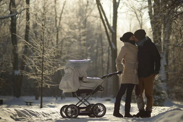 Jovem beijando e segurando pais com carrinho de bebê. Ensolarado parque de inverno nevado, árvores geladas geladas, creche e amor — Fotografia de Stock